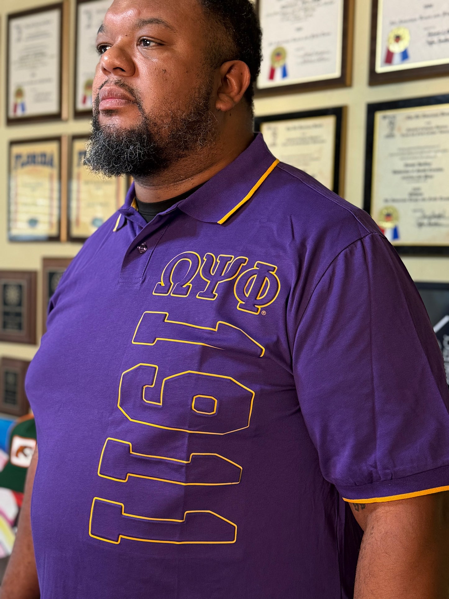 Man wearing a purple shirt with 'Omega Psi Phi' letters and number '1911' in front of framed certificates.